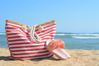 Photo of Stylish striped bag with slippers on sandy beach near sea. Space for text