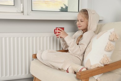 Photo of Little girl with cup of hot drink near heating radiator indoors