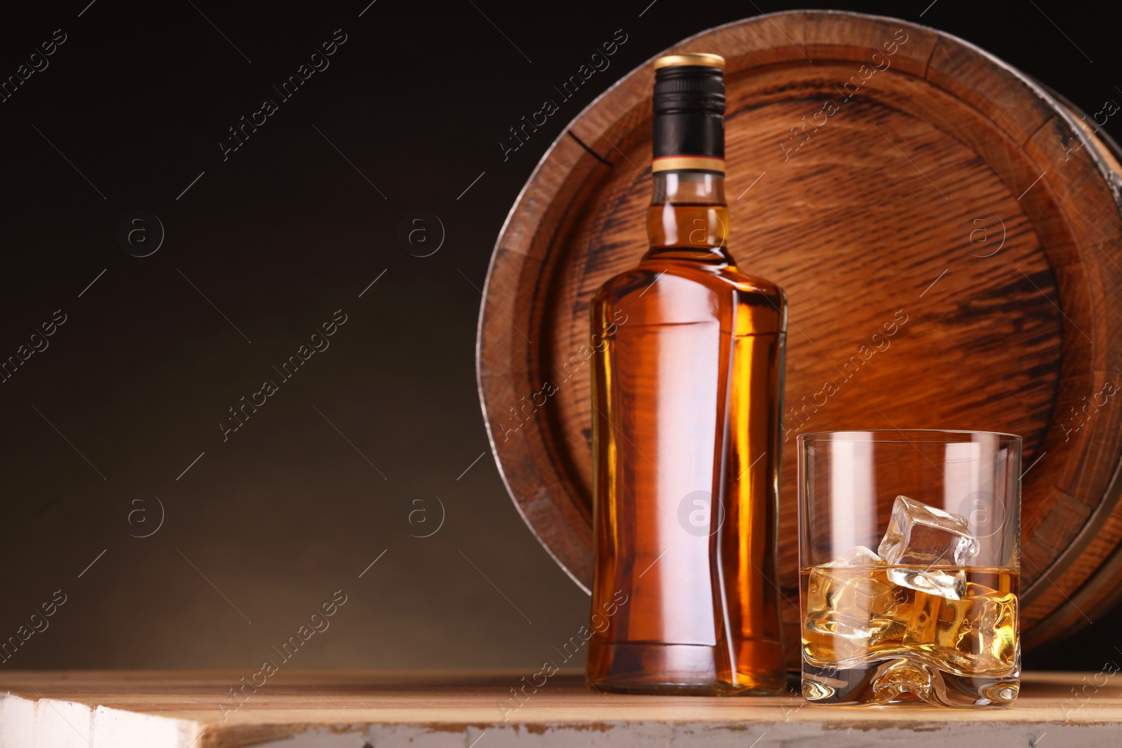 Photo of Whiskey with ice cubes in glass, bottle and barrel on wooden table against dark background, space for text