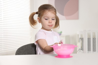 Cute little child eating tasty yogurt from plastic bowl with spoon at white table indoors