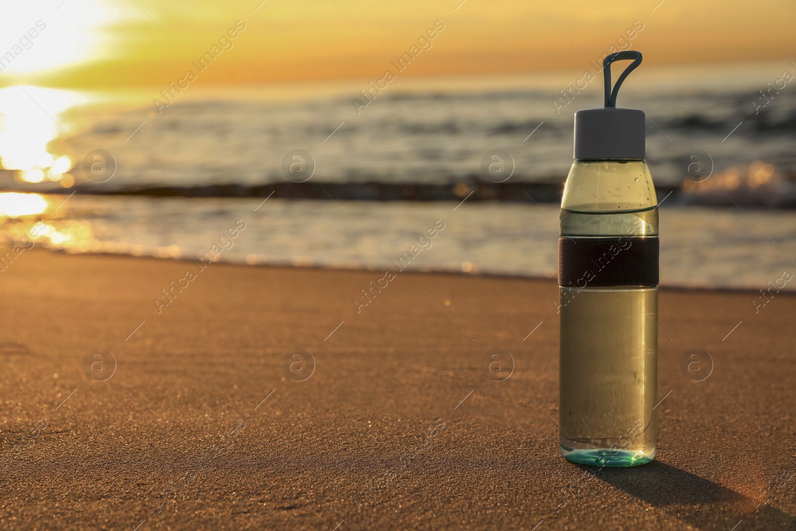 Photo of Glass bottle with water on wet sand near sea at sunset. Space for text