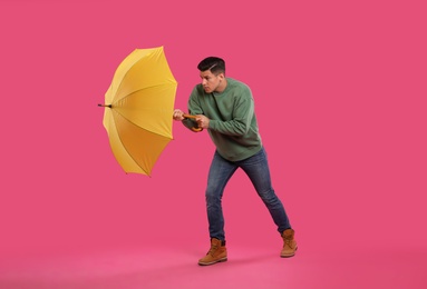 Emotional man with umbrella caught in gust of wind on pink background