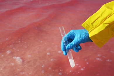 Photo of Laboratory worker with test tube taking sample from pink lake for analysis, closeup