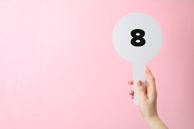Woman holding auction paddle with number 8 on pink background, closeup. Space for text