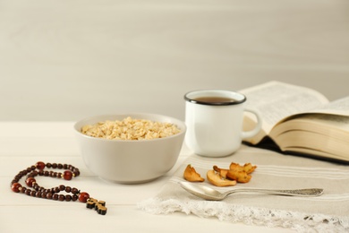 Photo of Rosary beads, oatmeal porridge, dried apples, drink and Bible on white wooden table. Lent season