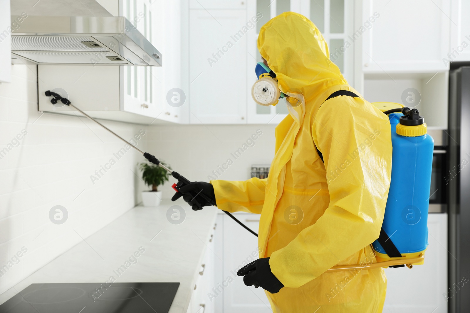 Photo of Pest control worker in protective suit spraying insecticide on furniture indoors