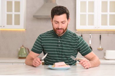 Photo of Happy man holding knife and fork with sausage at table in kitchen