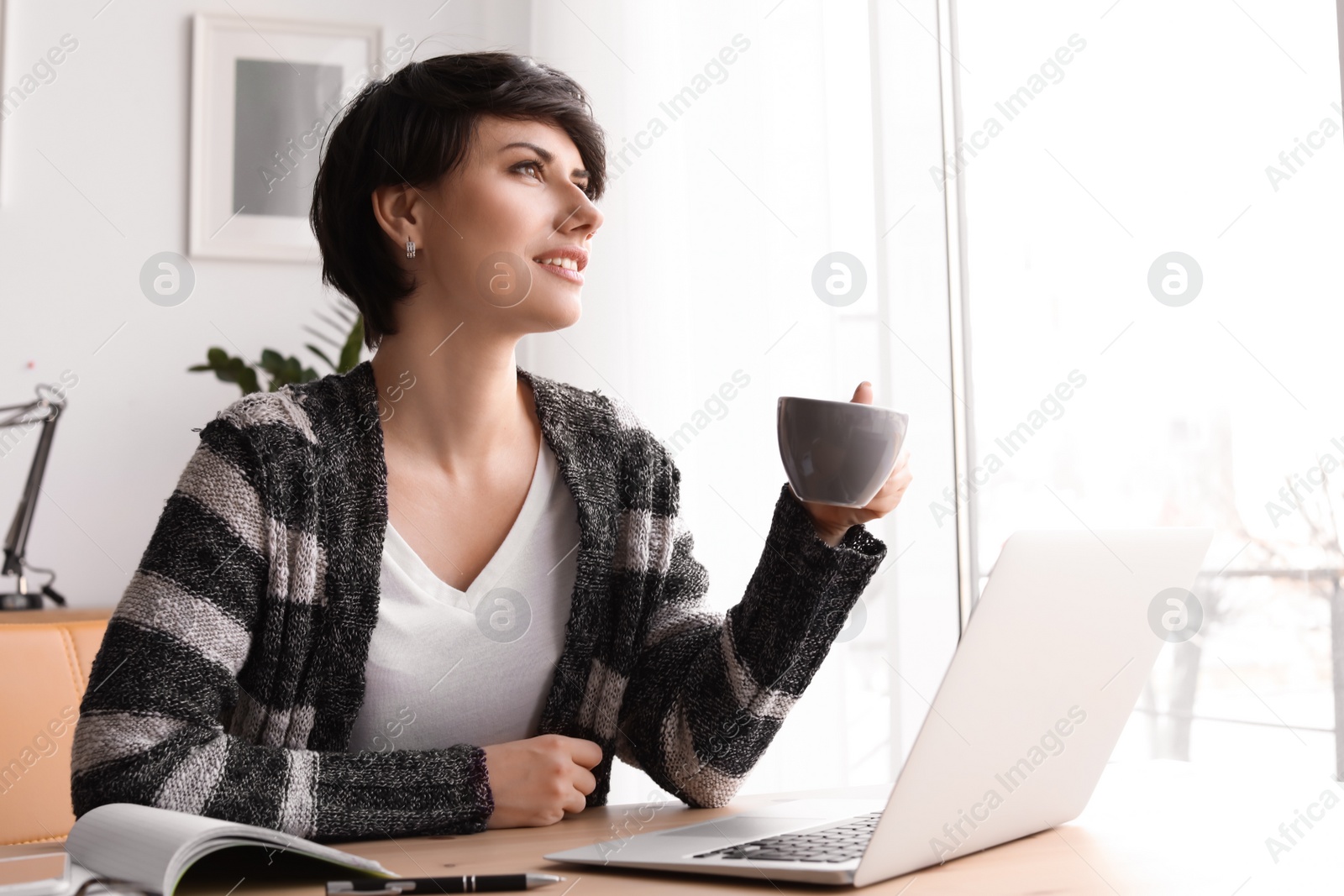 Photo of Young woman drinking coffee while working with laptop at desk. Home office