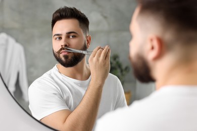 Photo of Handsome young man trimming mustache with scissors near mirror in bathroom