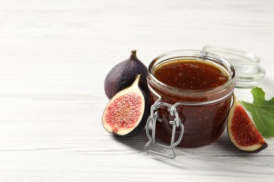 Photo of Glass jar with tasty sweet jam, green leaf and fresh figs on white wooden table. Space for text