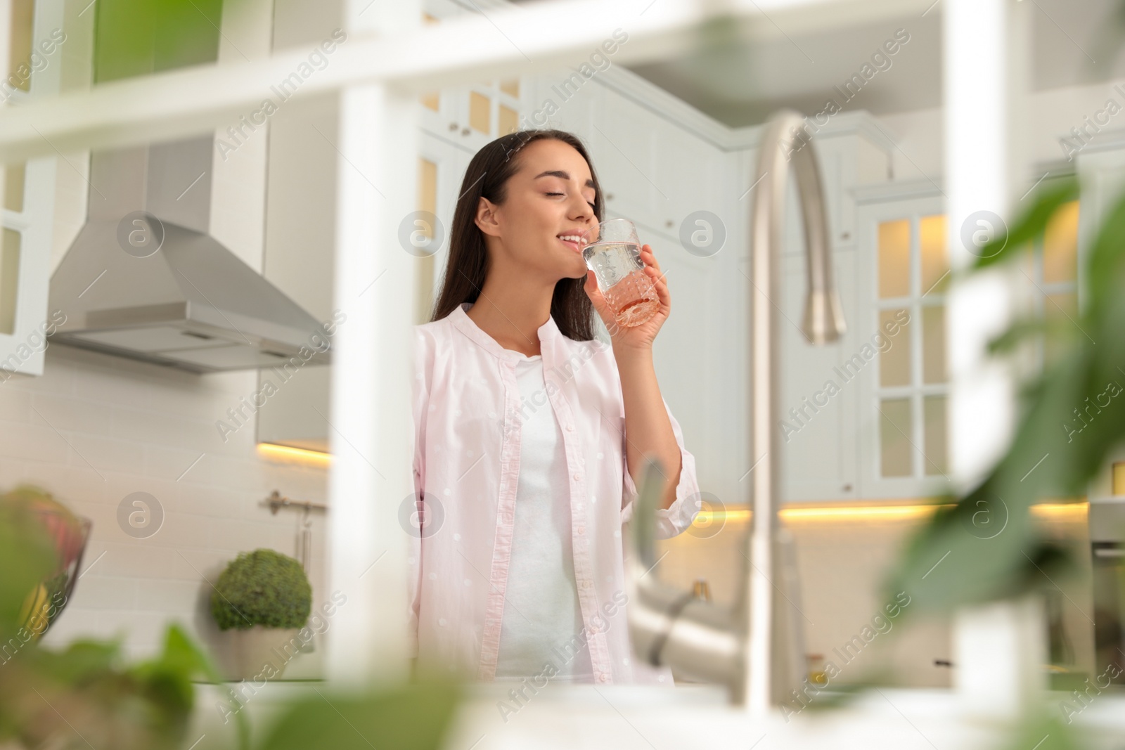 Photo of Young woman drinking pure water from glass in kitchen, view through window