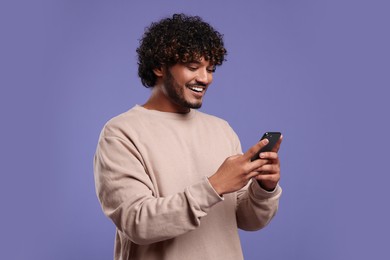 Handsome smiling man using smartphone on violet background, space for text