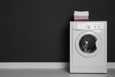 Photo of Modern washing machine with stack of towels near black wall, space for text. Laundry day