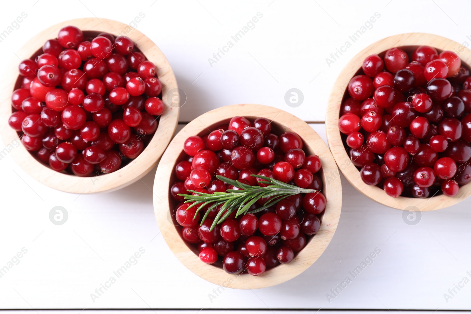 Photo of Fresh ripe cranberries in bowls and rosemary on white wooden table, top view