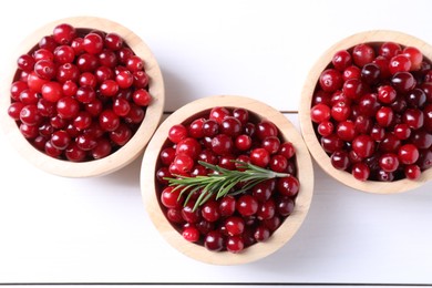 Fresh ripe cranberries in bowls and rosemary on white wooden table, top view