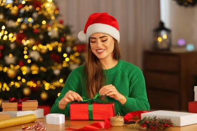 Beautiful young woman in Santa hat making Christmas gift at table in room