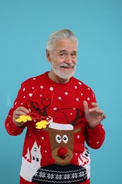 Photo of Happy senior man in Christmas sweater holding funny glasses on light blue background