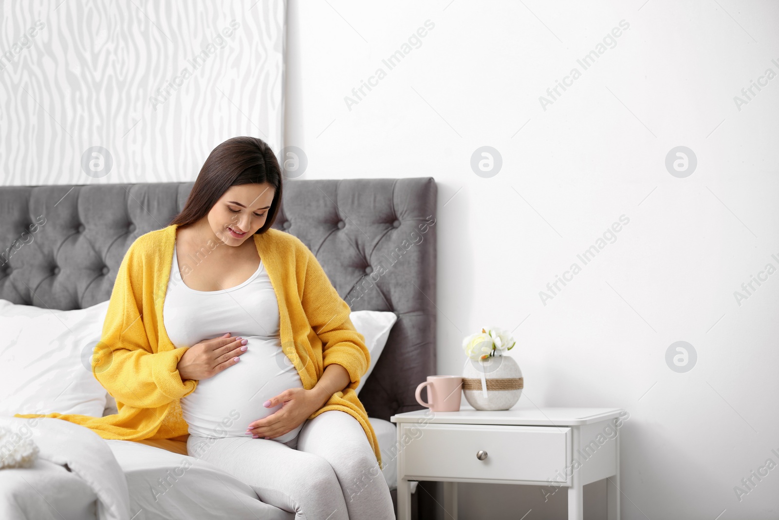 Photo of Young beautiful pregnant woman sitting on bed and touching her belly at home