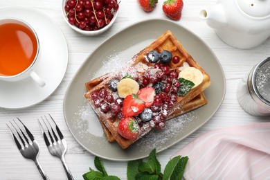 Photo of Plate of delicious Belgian waffles with berries, banana and tea on white wooden table, flat lay