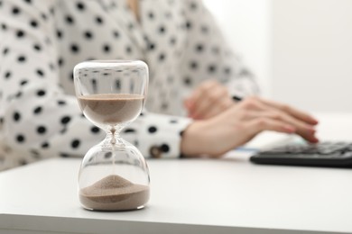 Photo of Hourglass with flowing sand on desk. Woman taking notes while using calculator indoors, selective focus