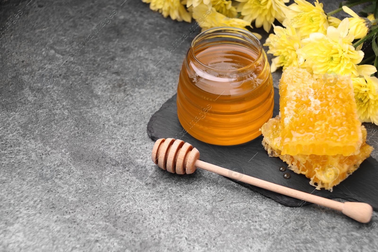 Photo of Sweet golden honey in jar, dipper, chrysanthemum flowers and pieces of honeycomb on grey textured table, space for text