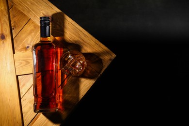 Whiskey with ice cubes in glass and bottle on wooden crate against black background, top view. Space for text