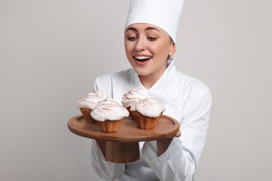 Photo of Happy professional confectioner in uniform holding delicious cupcakes on light grey background