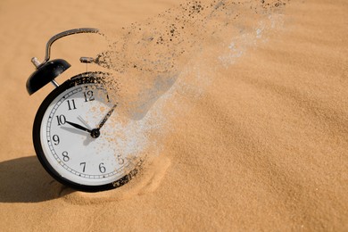Image of Time is running out. Black alarm clock vanishing on sand in desert