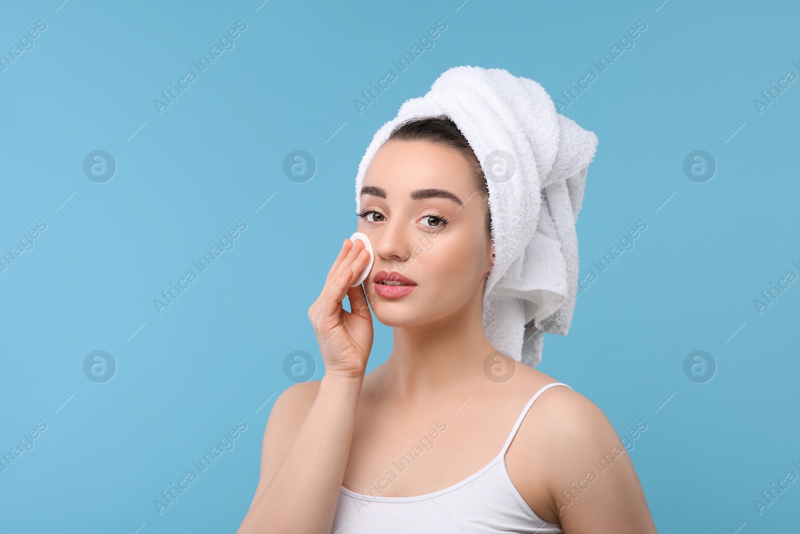 Photo of Beautiful woman in terry towel removing makeup with cotton pad on light blue background, space for text