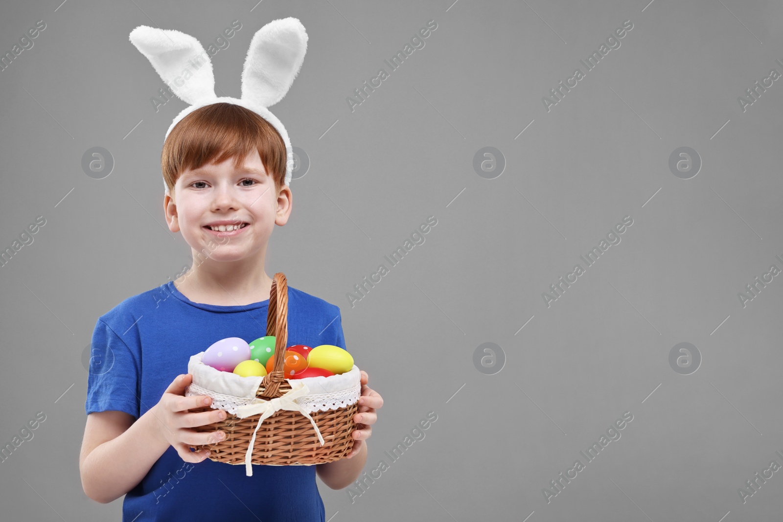 Photo of Easter celebration. Cute little boy with bunny ears and wicker basket full of painted eggs on grey background. Space for text