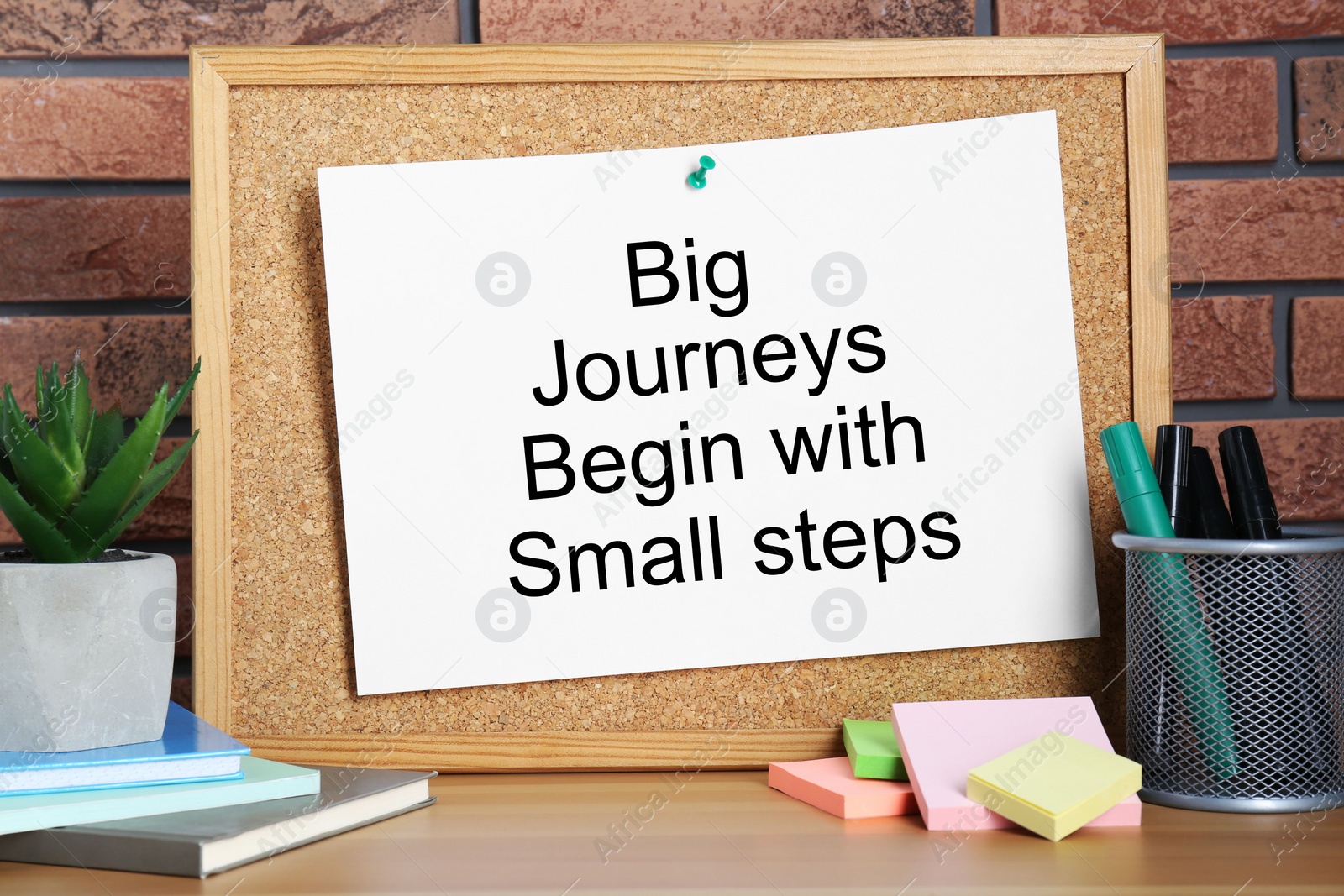 Photo of Corkboard and phrase Big Journeys Begin With Small Steps, houseplant, markers and books on table against brick wall. Motivational quote