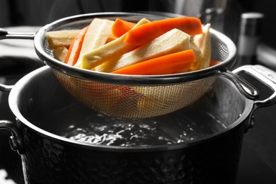 Photo of Sieve with cut parsnips and carrots over pot of boiling water in kitchen, closeup