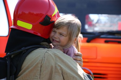 Photo of Firefighter in uniform holding rescued little girl near fire truck outdoors. Save life