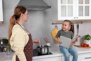 Photo of Happy woman and her daughter reading recipe in culinary magazine while cooking at home