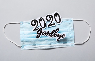 Photo of Text Goodbye 2020 and medical face mask on light grey background, flat lay