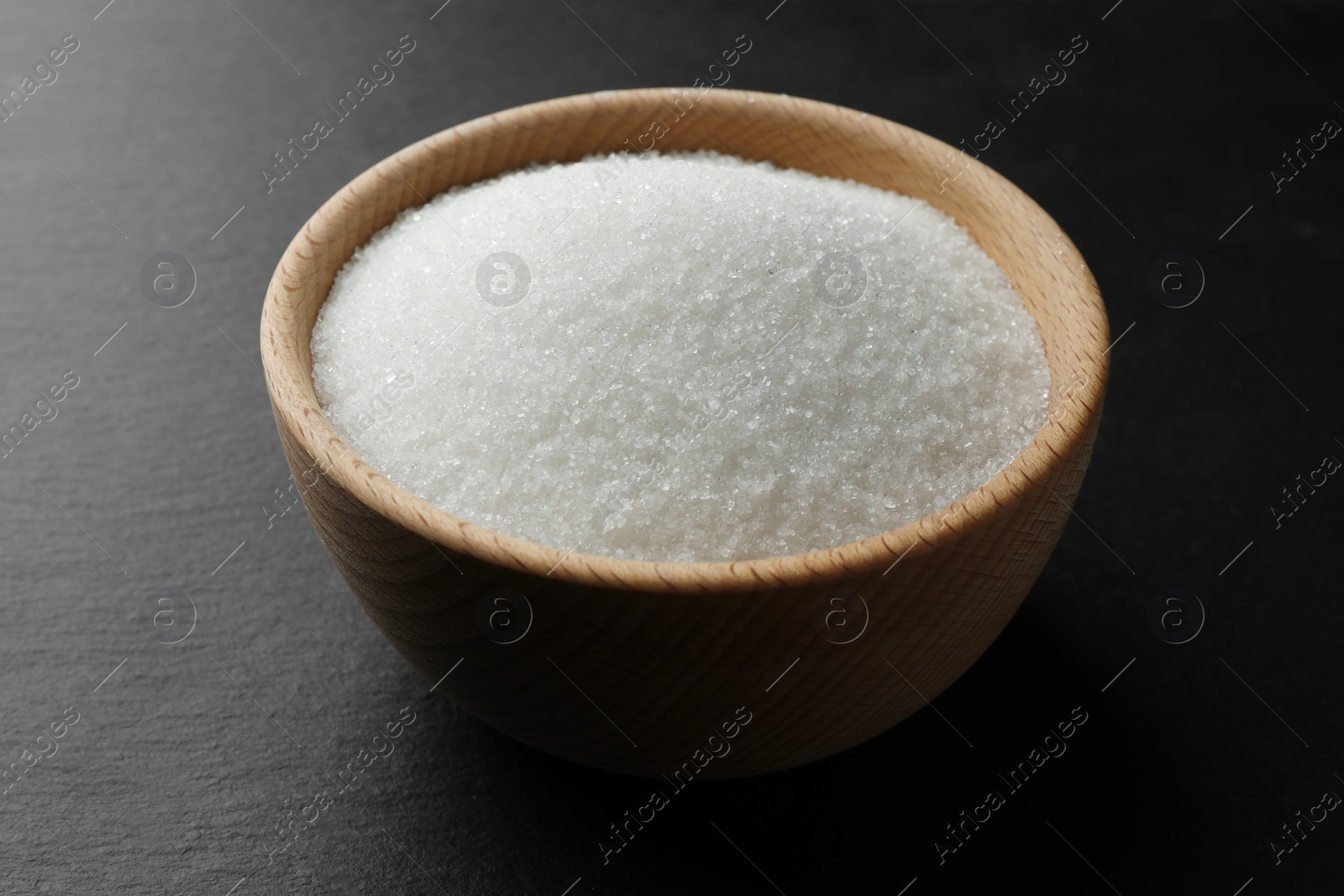 Photo of Granulated sugar in bowl on black table, closeup