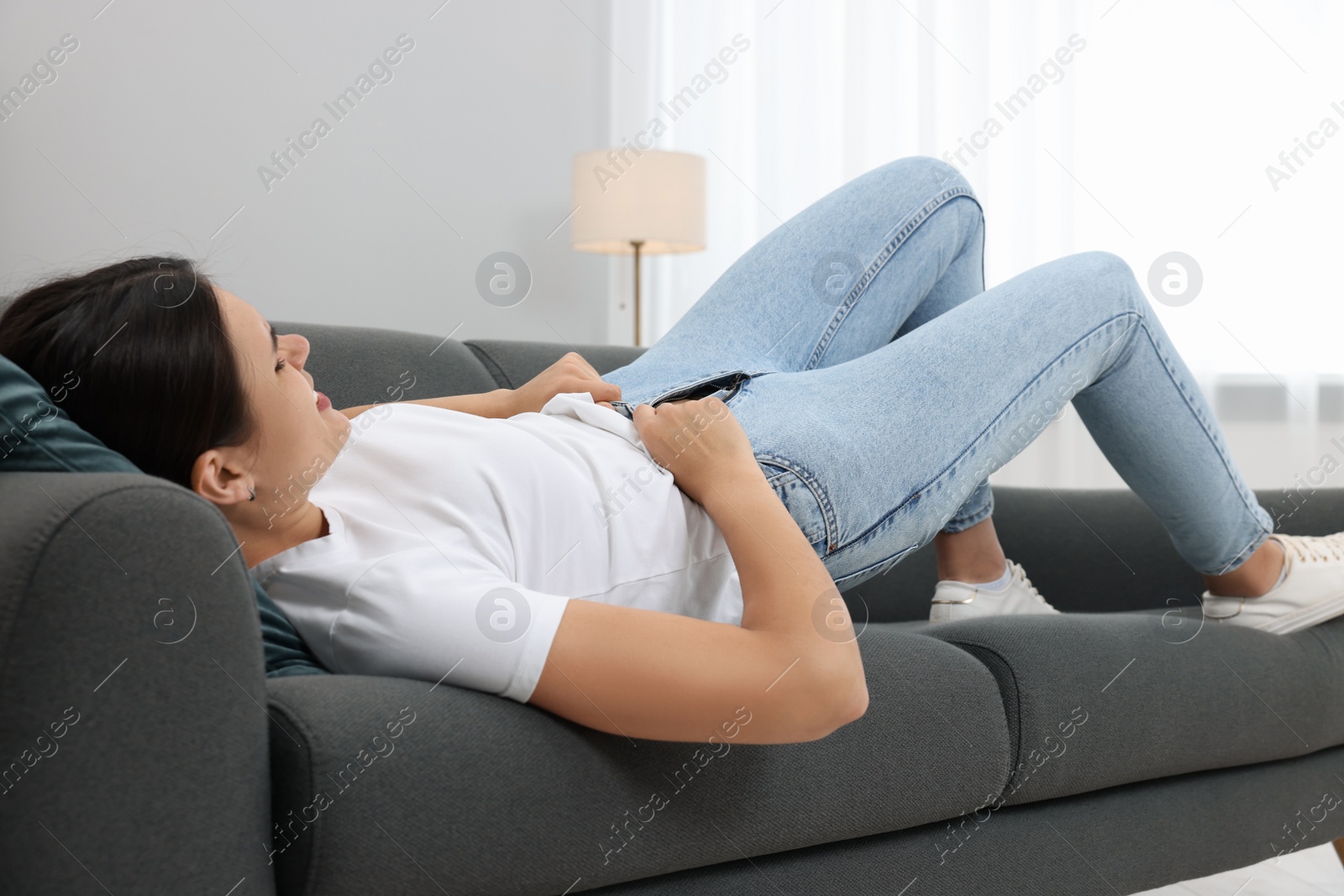 Photo of Young woman struggling to squeeze into tight jeans while lying on sofa at home