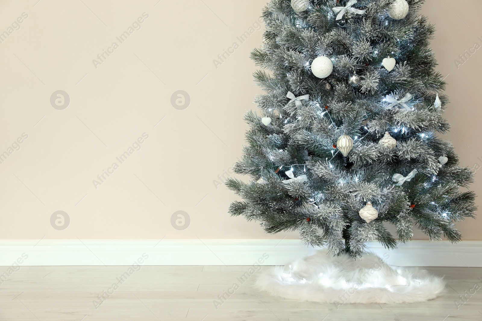 Photo of Decorated Christmas tree with white faux fur skirt near beige wall. Space for text
