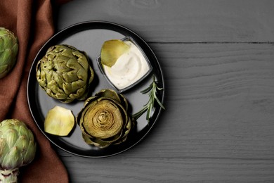 Delicious cooked artichokes with tasty sauce served on grey wooden table, flat lay. Space for text