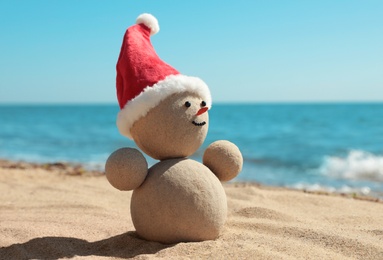 Photo of Snowman made of sand with Santa hat on beach near sea, closeup. Christmas vacation