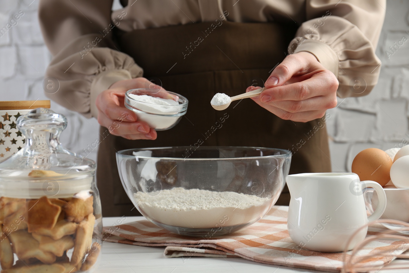 Photo of Woman with spoon and bowl of baking powder at white table, closeup