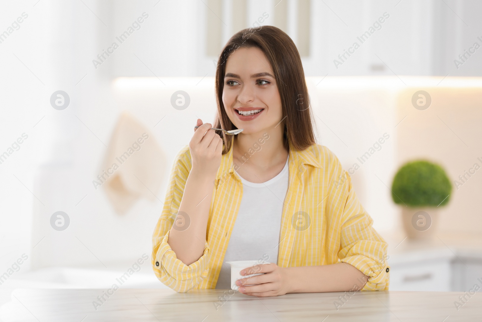 Photo of Young attractive woman eating tasty yogurt at table in kitchen