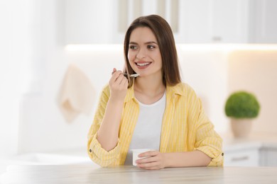 Photo of Young attractive woman eating tasty yogurt at table in kitchen