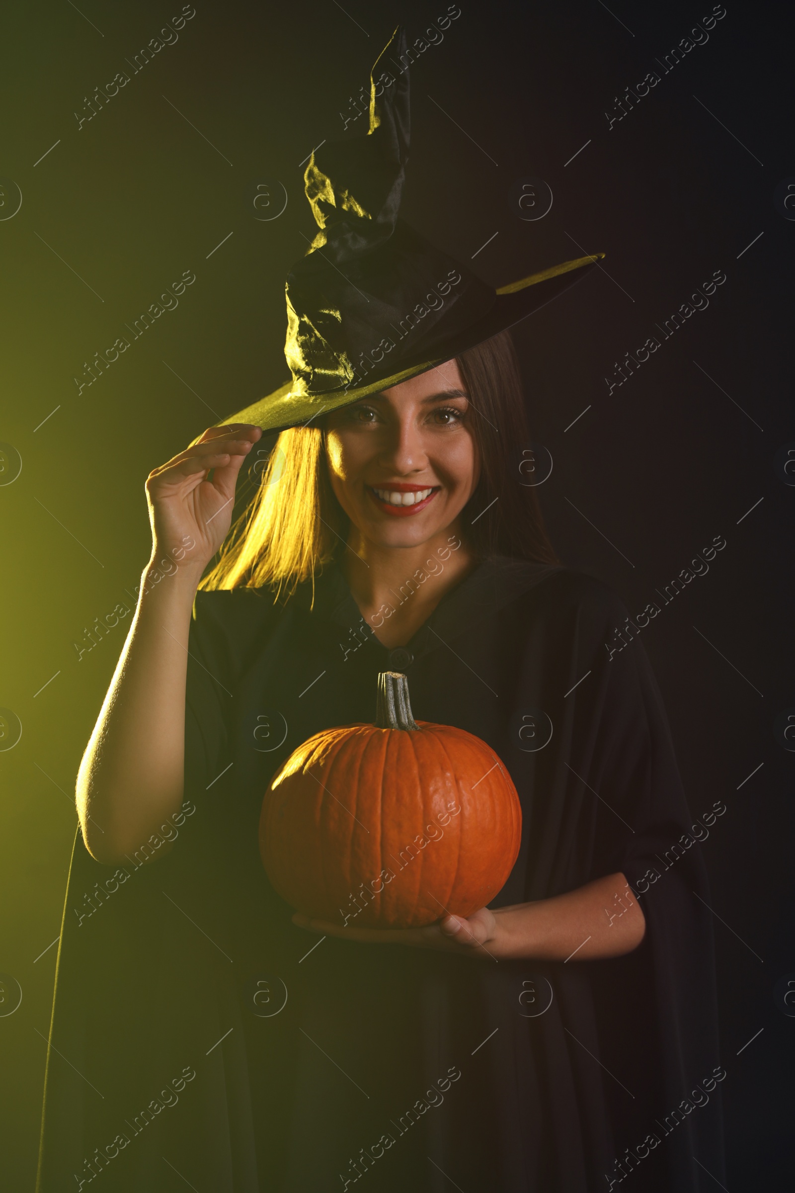 Photo of Young woman wearing witch costume with pumpkin on dark background. Halloween party