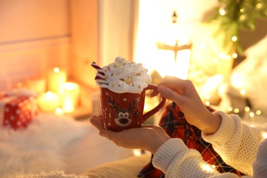 Photo of Woman holding cup of delicious drink with whipped cream indoors, closeup. Christmas celebration