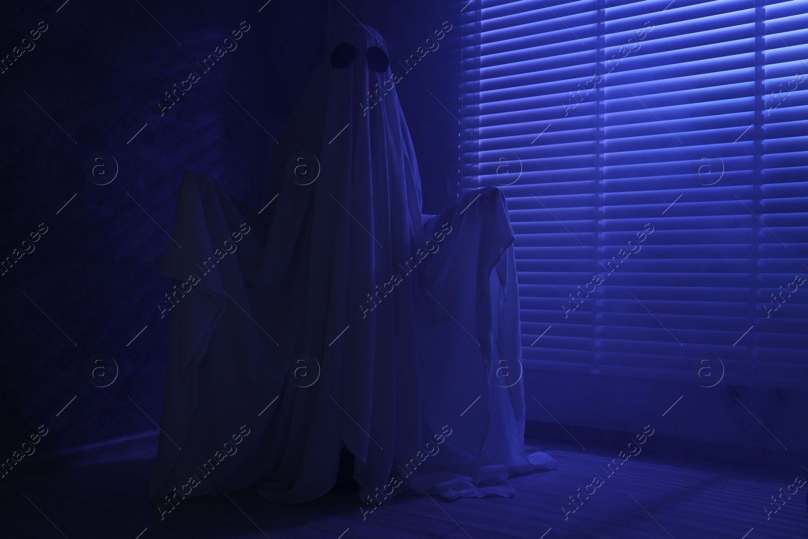 Photo of Creepy ghost. Woman covered with sheet near window in blue light