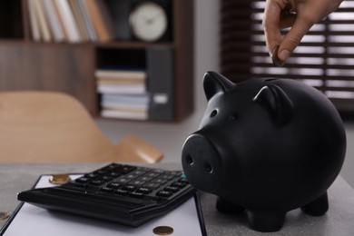 Woman putting coin into piggy bank at grey table indoors, closeup