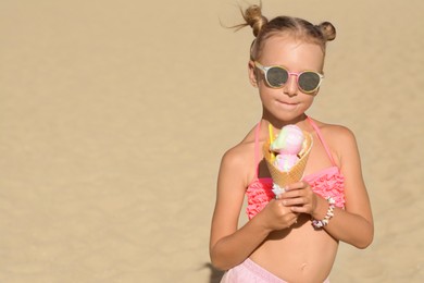 Photo of Adorable little girl in swimsuit with delicious ice cream at beach on sunny summer day, space for text