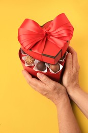 Photo of Woman with heart shaped box of delicious chocolate candies on yellow background, closeup