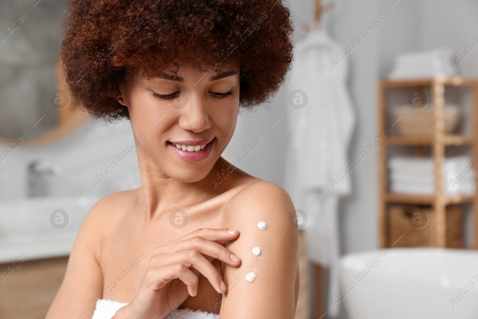Photo of Beautiful young woman applying body cream onto arm in bathroom, space for text